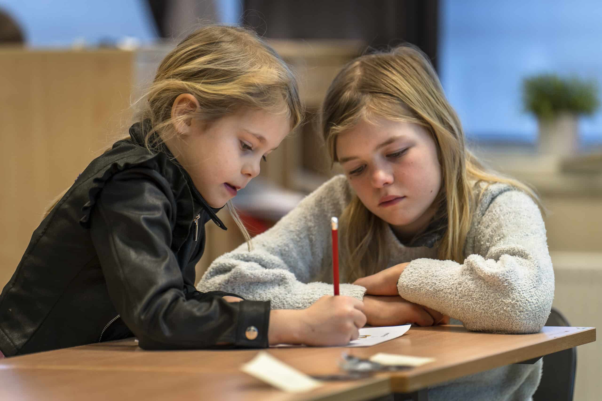 To jenter sitter ved siden av hverandre ved en pult. Hun ene holder en blyant og skriver på et ark, mens hun andre ser på. De bøyer seg over arket og ser veldig konsentrerte ut.